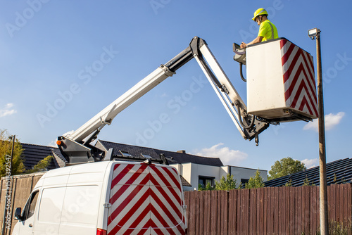 A technician in a bright vest works at height using a hydraulic boom lift truck near private homes. Concept of occupational safety and technical work.