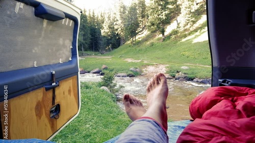 Waking up in a car with a view of a picturesque mountain meadow with rocks, a mountain river and a forest in the morning. Women legs in a camper sleeping bag against the backdrop of nature