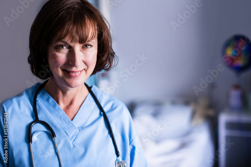 Smiling female nurse standing in patient room in blue scrubs with stethoscope, copy space