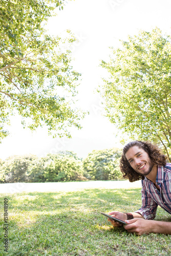 Man reclining on grassy lawn using tablet under leafy tree shade with sunlight at public park