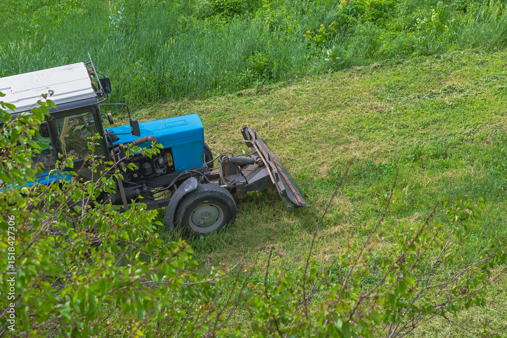 Obraz premium A tractor is driving across a green field with mowed grass, the tractor is clearing overgrown green grass, tractor work on a green field, agricultural work.