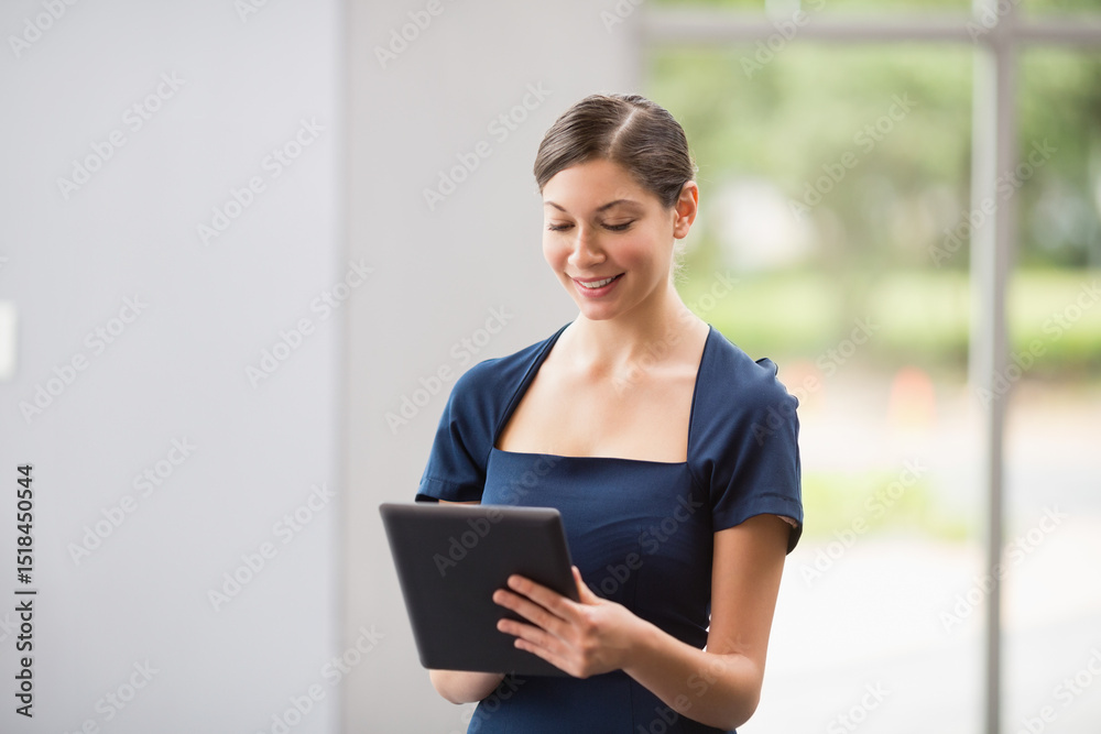 Fototapeta premium Woman standing in office lobby wearing navy blue dress holding black tablet and scrolling by window