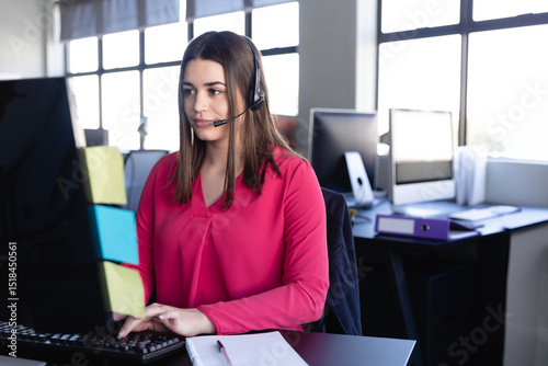 Woman call center agent wearing pink blouse typing on keyboard at office with headset, sticky notes