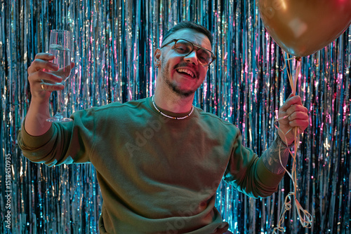 Portrait of young adult Caucasian man smiling at camera holding champagne glass and gold balloons celebrating birthday against shiny party backdrop photo
