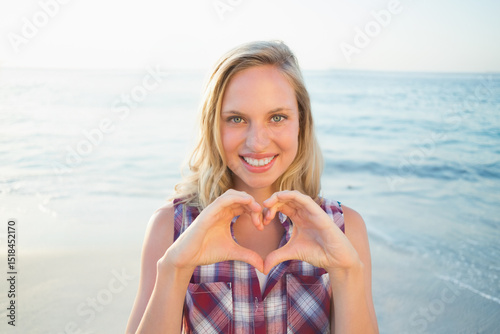 Woman smiling, making heart shape with hands at beach shoreline wearing button-up shirt