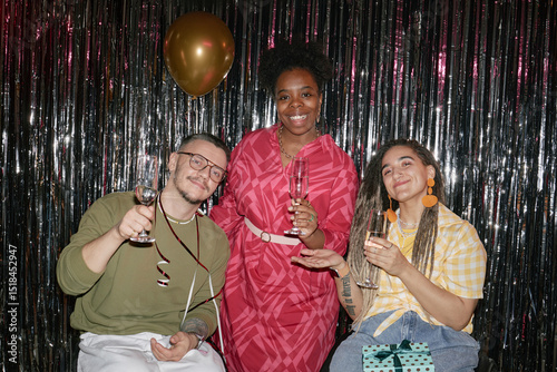 Multiethnic group of young adults celebrating birthday, smiling and holding glasses in front of metallic backdrop, Caucasian man with disability in wheelchair joining Black woman and Caucasian woman photo