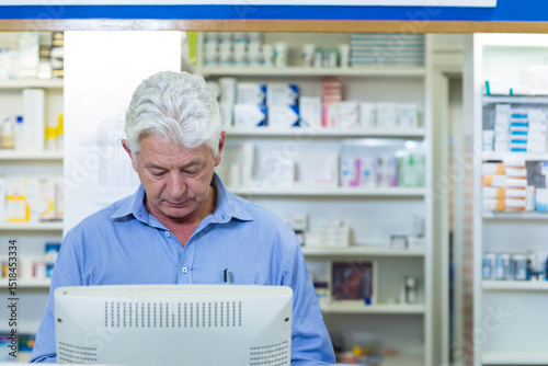 Senior male pharmacist in blue shirt checking white monitor at pharmacy counter and shelves