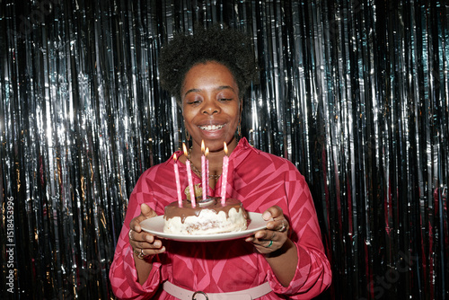 Portrait of adult Black woman smiling while holding birthday cake with lit candles in front of metallic backdrop, celebrating birthday and looking at cake photo