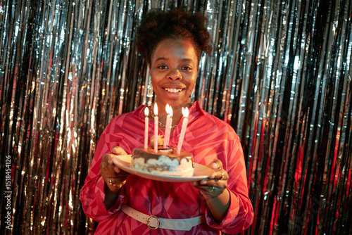 Portrait of young adult Black woman smiling and holding birthday cake with lit candles in front of metallic backdrop celebrating birthday in festive setting photo