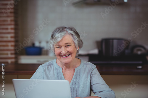 Senior woman typing on laptop at kitchen counter with blue pot and tiled backsplash