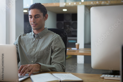 Middle Eastern man typing on laptop with notebook, coffee cup and speakers in office, copy space