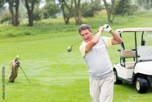 Senior man swinging driver golf club on fairway in polo shirt near golf cart, copy space