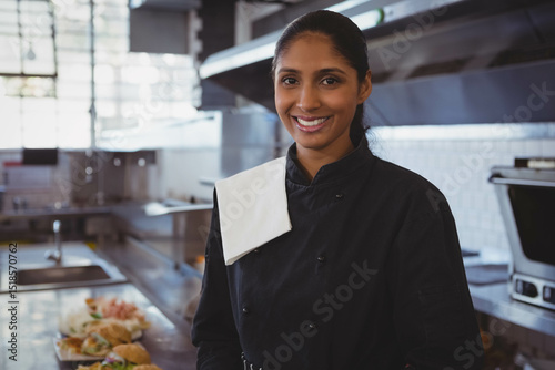 Indian female chef wearing black jacket smiling in kitchen by prep counter with plated sandwiches