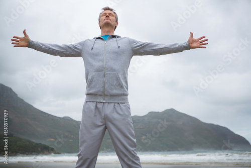 Middle-aged man in grey hoodie and sweatpants stretching arms and inhaling ocean breeze on beach