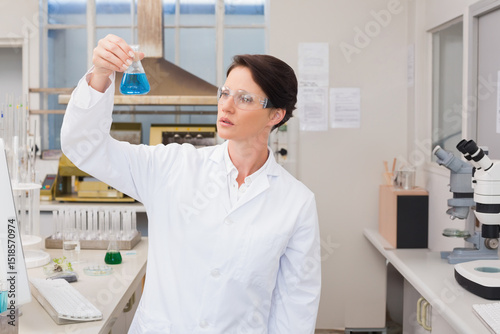 Female scientist holding up conical flask in lab inspecting blue solution wearing goggles