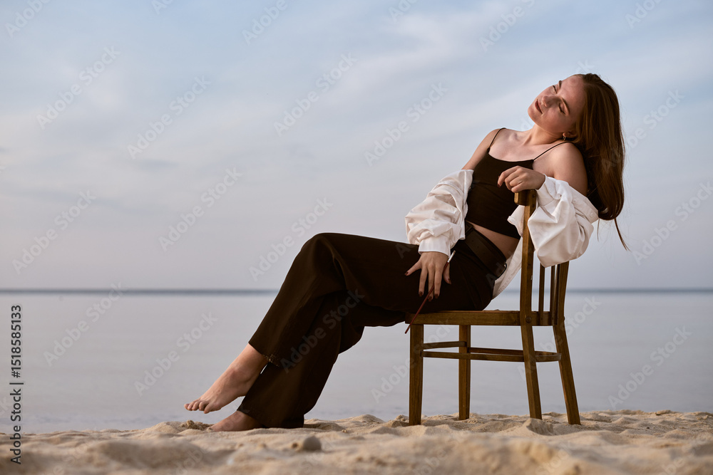 Fototapeta premium Young female painter sits thoughtfully on chair by tranquil beach. Serene setting and soft lighting evoke creativity and introspection, symbolizing artistic inspiration