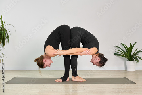 Two adult women perform synchronized yoga pose on mat. Dressed in black, they balance skillfully, adding symmetry to serene, minimalist room