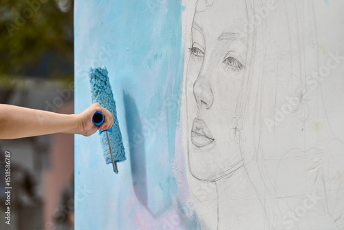 Hand applies soft blue paint over pencil sketch of young female face on white wall. Natural outdoor setting, gentle lighting, artistic enhancement in progress