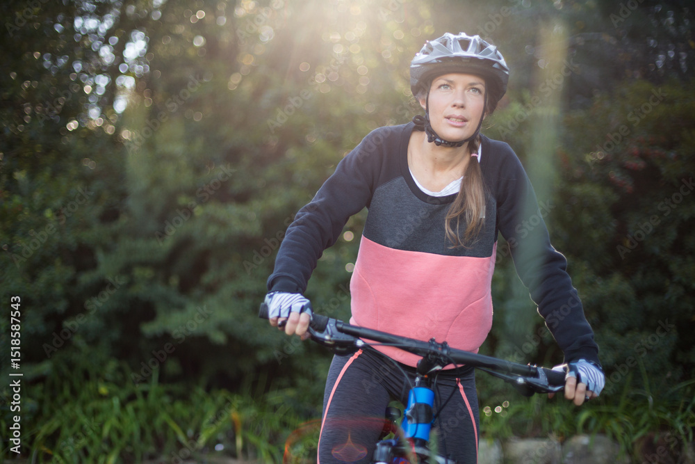 Obraz premium Woman cyclist riding mountain bike along wooded trail in sunlight wearing helmet, fingerless gloves