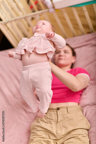 Young brunette woman in pink shirt gently lifts baby girl in pale pink outfit. Infant shows curiosity cozy setting with soft lighting and pastel tones