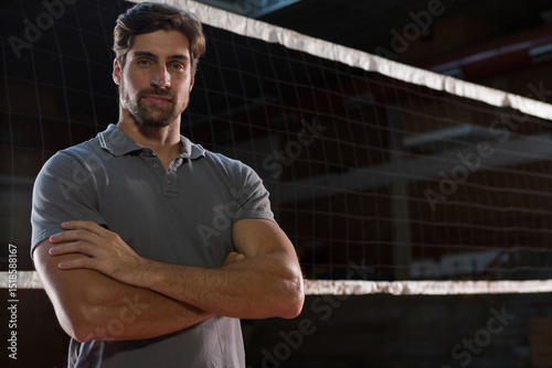 Man standing with arms crossed wearing grey polo shirt at volleyball net posts in sports hall