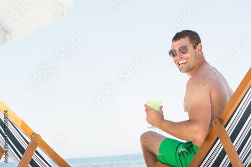 Man in swim trunks relaxing on chair under umbrella holding martini glass on beach, copy space
