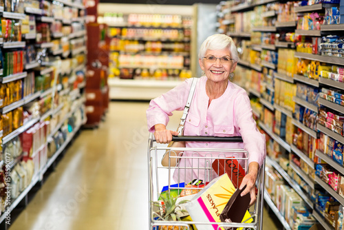 Senior female customer pushing shopping cart down grocery aisle, carrying cereal box and pineapple