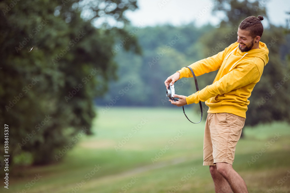 Fototapeta premium Man wearing yellow hoodie and tan shorts holding film camera smiling in grassy meadow, copy space