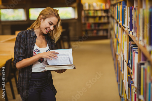 Woman in her twenties holding two books flipping one open beside bookshelf at library, copy space