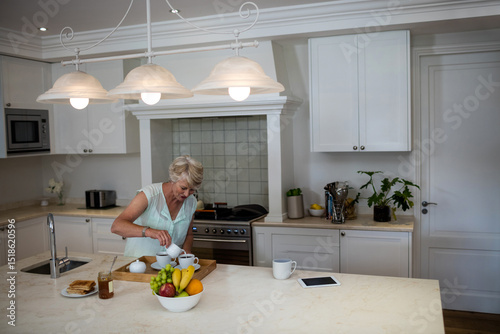 Senior woman pouring tea at marble kitchen island with wooden tray teapot cups honey jar
