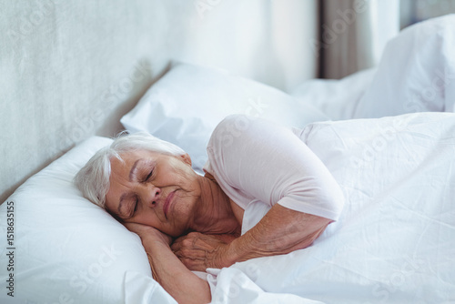 Senior woman lying on side in bed wearing sleep top and hugging white pillow against headboard