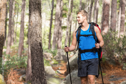 Man hiking rocky trail in pine forest gripping trekking poles and carrying backpack, copy space