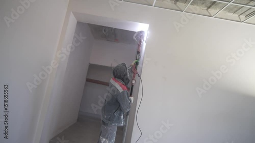 Worker sands wall removing plaster flaws with care. Dust swirls in dim room under steady hands. Man evens out plaster perfecting each patch