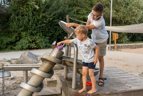 Happy brothers playing with Archimedes screw water system at outdoor playground on warm summer day. Concept of teamwork, child development, interactive science, and educational fun in public park.