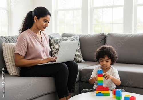 Busy indian mother using laptop and working remotely on sofa while her little son is playing with colorful building blocks in living room