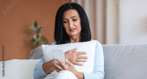 Free Download Remote Distant Communication. Senior woman with dark long hair holding mobile phone, making online video call and talking to children or watching video, embracing pillow, sitting on sofa at home - GFXLoad