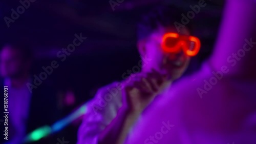 Close up of young man in red LED glasses dances energetically at a vibrant nightclub, surrounded by neon lights and partygoers. Concept of nightlife, clubbing, and celebration themes, college party