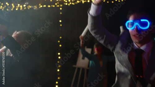 Close up of groom in a stylish suit dances joyfully at his wedding party, wearing glowing blue LED glasses. Concept of festive atmosphere, happy memories of celebrate college night party