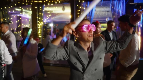 Stylish bride and groom wearing pink LED glasses dances enthusiastically at a wedding party, holding a glow stick. Concept of event, celebration, and nightlife themes, many people dance