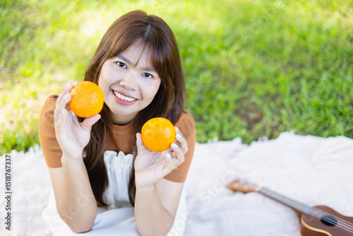 Young woman enjoys picnic outdoors holding two oranges with guitar nearby, capturing leisure moment with people and family concept in natural setting