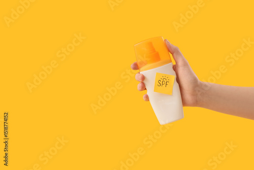 Woman holding bottle of sunscreen cream and sticky note with word SPF on yellow background