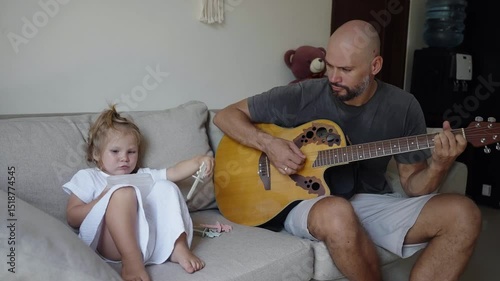 Dad plays the guitar for his small daughter sitting on sofa. Cozy family of father and his child in a modern home, education music, parenting. Happy memories of childhood and fatherhood