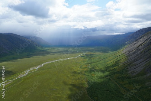 Vast green valley with winding river under cloudy sky and sun rays breaking through clouds over surrounding mountains