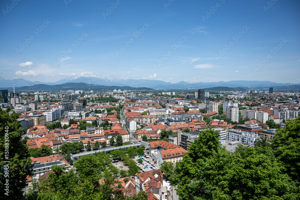 Fototapeta premium Panoramic view of Ljubljana city center featuring traditional architecture with terracotta roofs, modern buildings, and Julian Alps backdrop under clear blue sky