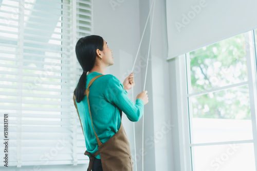 Asian housewife, wearing a green sweater and brown apron, adjusts the window blinds to let in natural light. Her attention to detail helps create a clean, bright, and welcoming home environment. Free Download Asian housewife, wearing a green sweater and brown apron, adjusts the window blinds to let in natural light. Her attention to detail helps create a clean, bright, and welcoming home environment. - GFXLoad