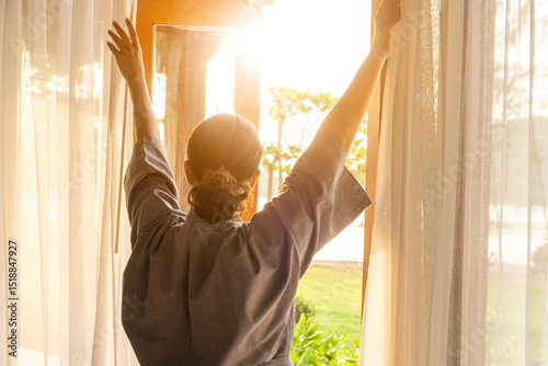 The woman was at the window in the bedroom. She opened the curtains on the window. In the morning and she looks at the view of mountains and trees at sunrise.	