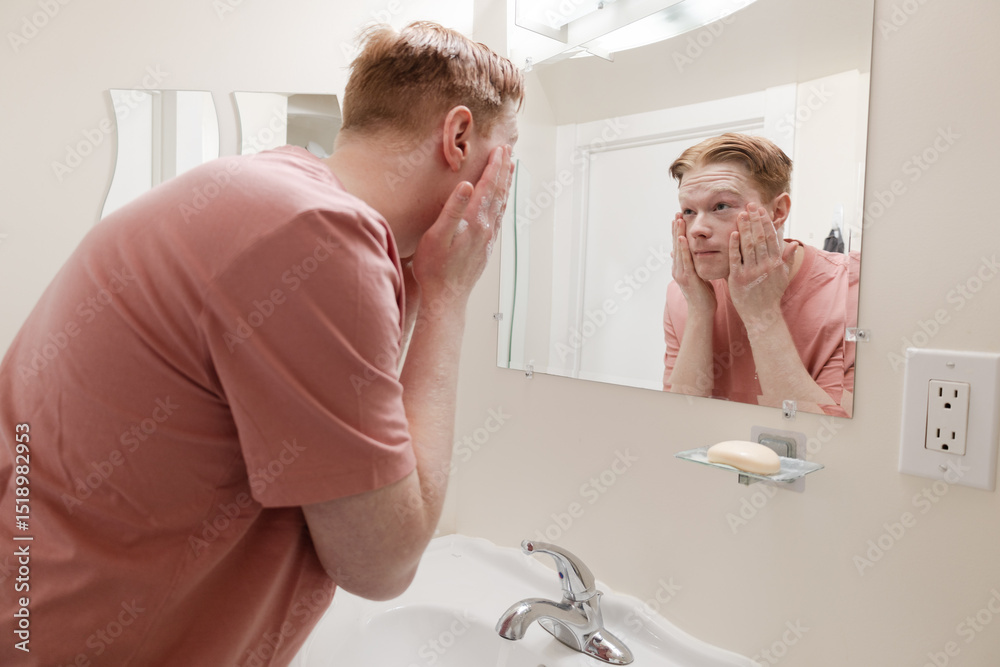 Fototapeta premium A Medium shot of a Man Washing Face at Bathroom Sink Candid image of a young man washing his face in a bathroom, captured through the mirror’s reflection. He is wearing a pink shirt.