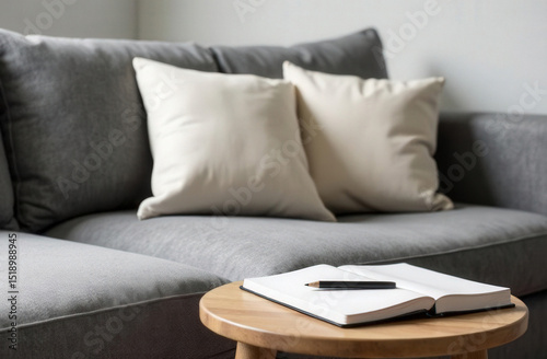 Monochrome interior vignette with grey sofa, off-white pillow and a minimal wooden table with an open blank notebook and a pencil, white wall in background, balanced composition