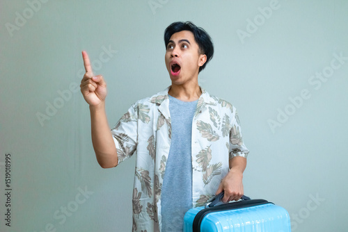Portrait of a young Asian man in summer clothes pointing to the side with a funny wow expression while carrying a suitcase, isolated on a clear background, travel concept