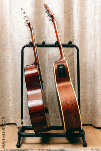 Two acoustic guitars stand side-by-side on a black metal guitar stand in front of beige curtains